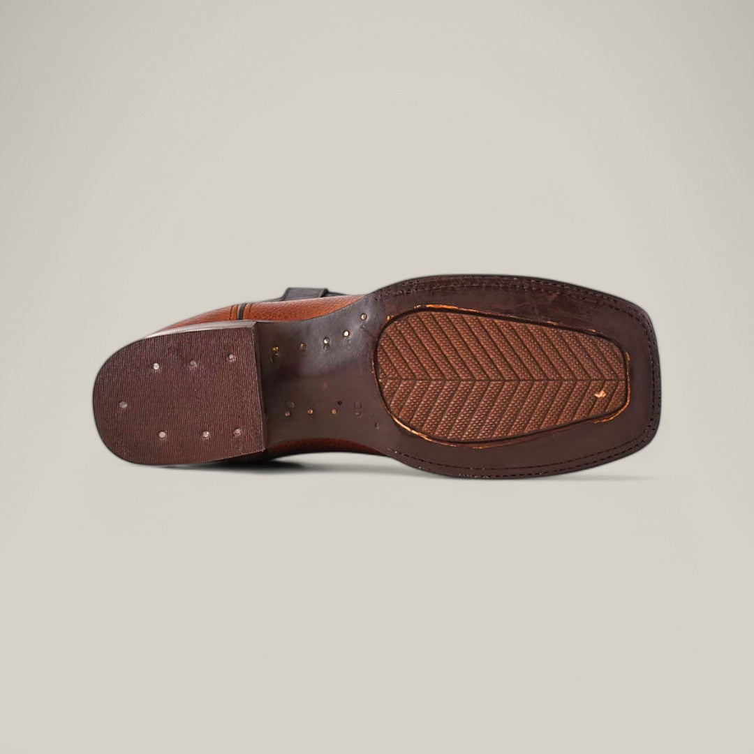 The image features the sole of a Grisly Tan - Square Toe boot, made of premium leather with stitching and small perforations. This Western-style shoe has a textured grip and rectangular heel, set against a plain light gray background.