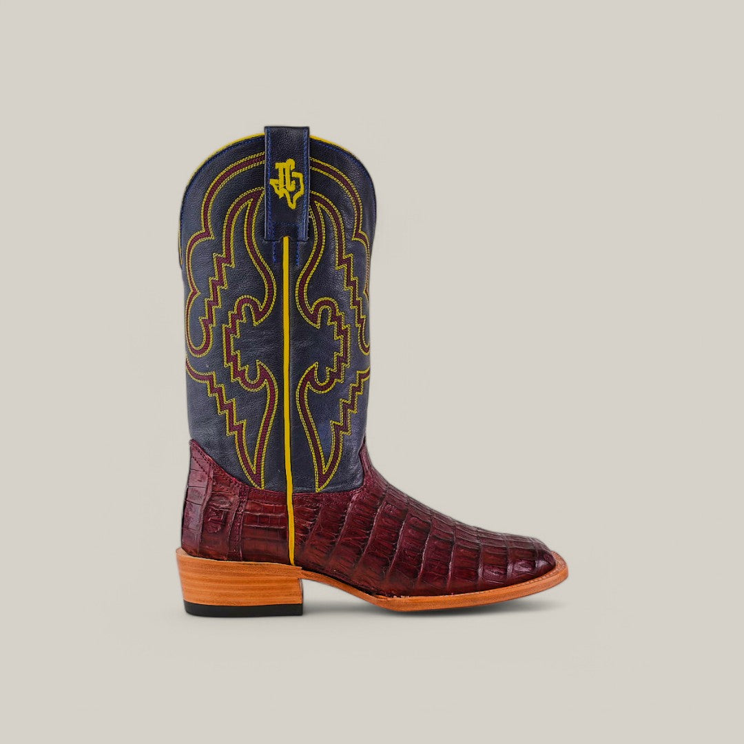The Exotic Caiman Belly Tail boot in wine features a maroon crocodile pattern, a navy shaft with yellow stitching and logo, square toe design, brown stacked heel, and pull strap on a light gray background.