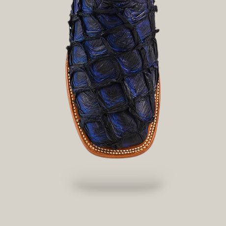 Close-up of the toe of an Exotic Pirarucu Fish - Navy Black boot with a square tip, distinct textured scales, brown trim, and decorative stitching, highlighting luxury footwear against a plain beige background.