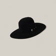 The 25X Grizzly - Black - TC Hats is a wide-brimmed black felt hat with an open, rounded crown and a decorative band with two small metal buckles, shown against a plain light beige background.
