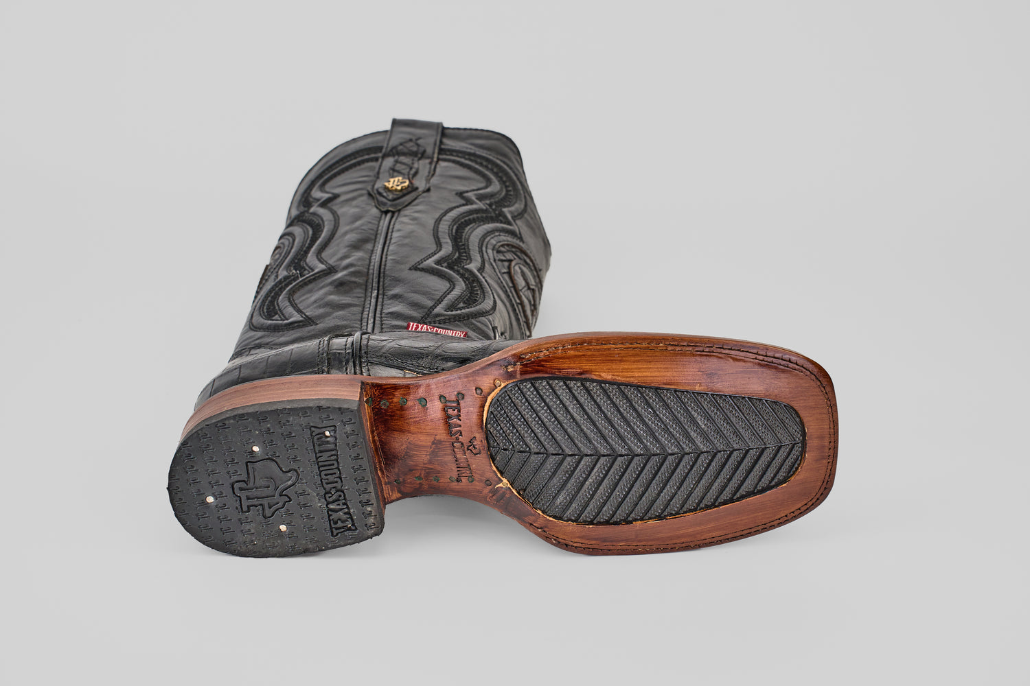 The Exotic Alligator Tail - Black - Square Toe Boot lies on its side, showcasing a wooden-finish sole with textured rubber grip and visible stitching, plus decorative upper stitching that highlights classic cowboy boot style.