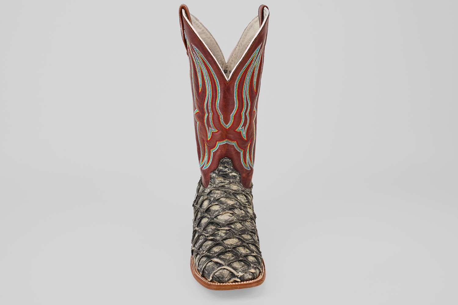 The Robinson - Newspaper - Square Toe is a single cowboy boot featuring a brown and red stitched leather upper and a textured, scaly dark brown Purarucu leather lower, displayed against a light gray background.