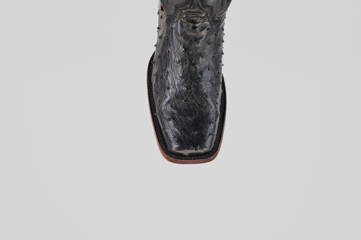 A close-up of the Exotic Ostrich - Black - Rodeo Toe boot shows its textured leather, square toe, and detailed stitching along the brown sole, exemplifying classic western style against a plain light gray background.