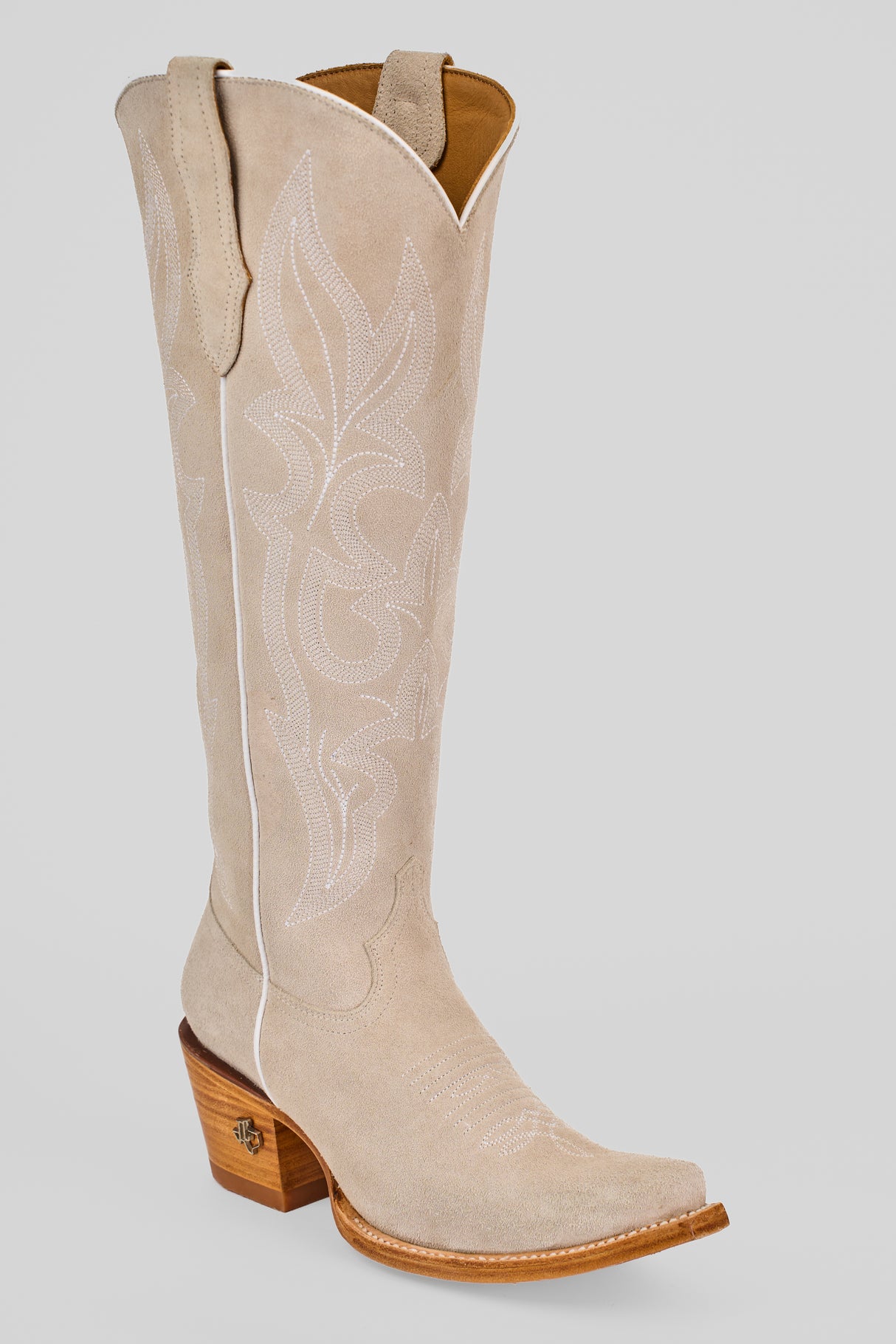 The Allison Ivory - Tall Shaft - Snip Toe boot features light beige suede, decorative white stitching, a pointed toe, wooden angled heel, and pull straps, shown against a plain light gray background.