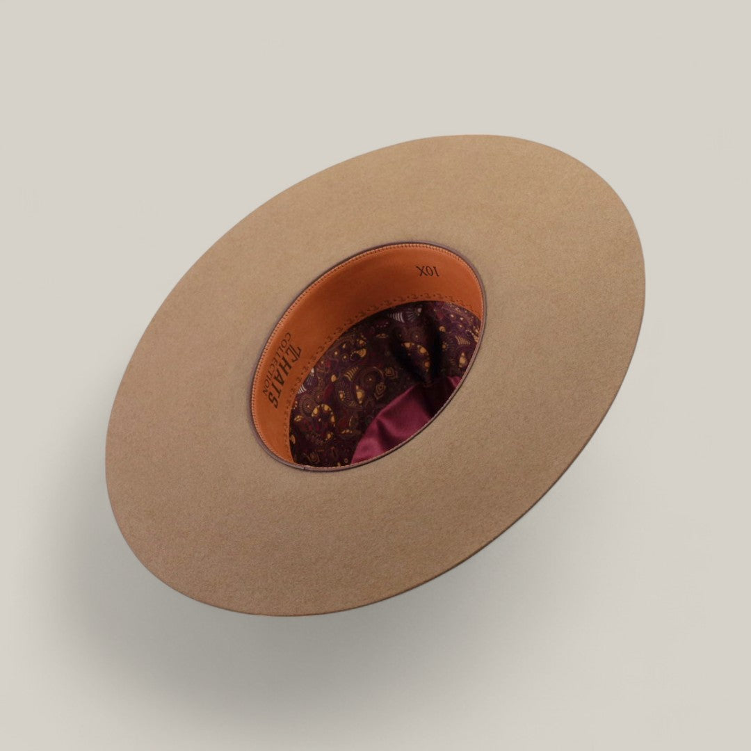 The 10x - Natural - TC Hats wide-brimmed open crown hat is pictured upside down, showing its brown patterned inner lining and tan leather sweatband with stamped writing, set against a plain beige background.