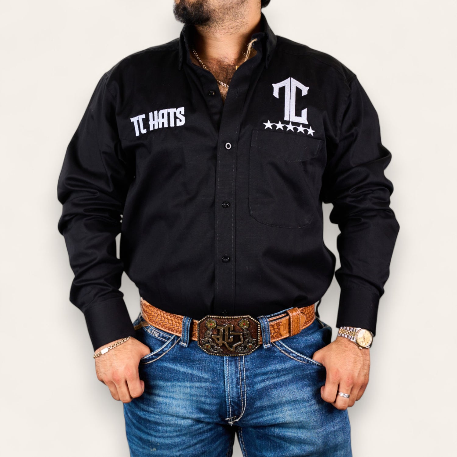 A man models the TC Hats - Black & White shirt with logo embroidery, paired with blue jeans, a brown tooled leather belt, gold watch, and jewelry against a plain white background.