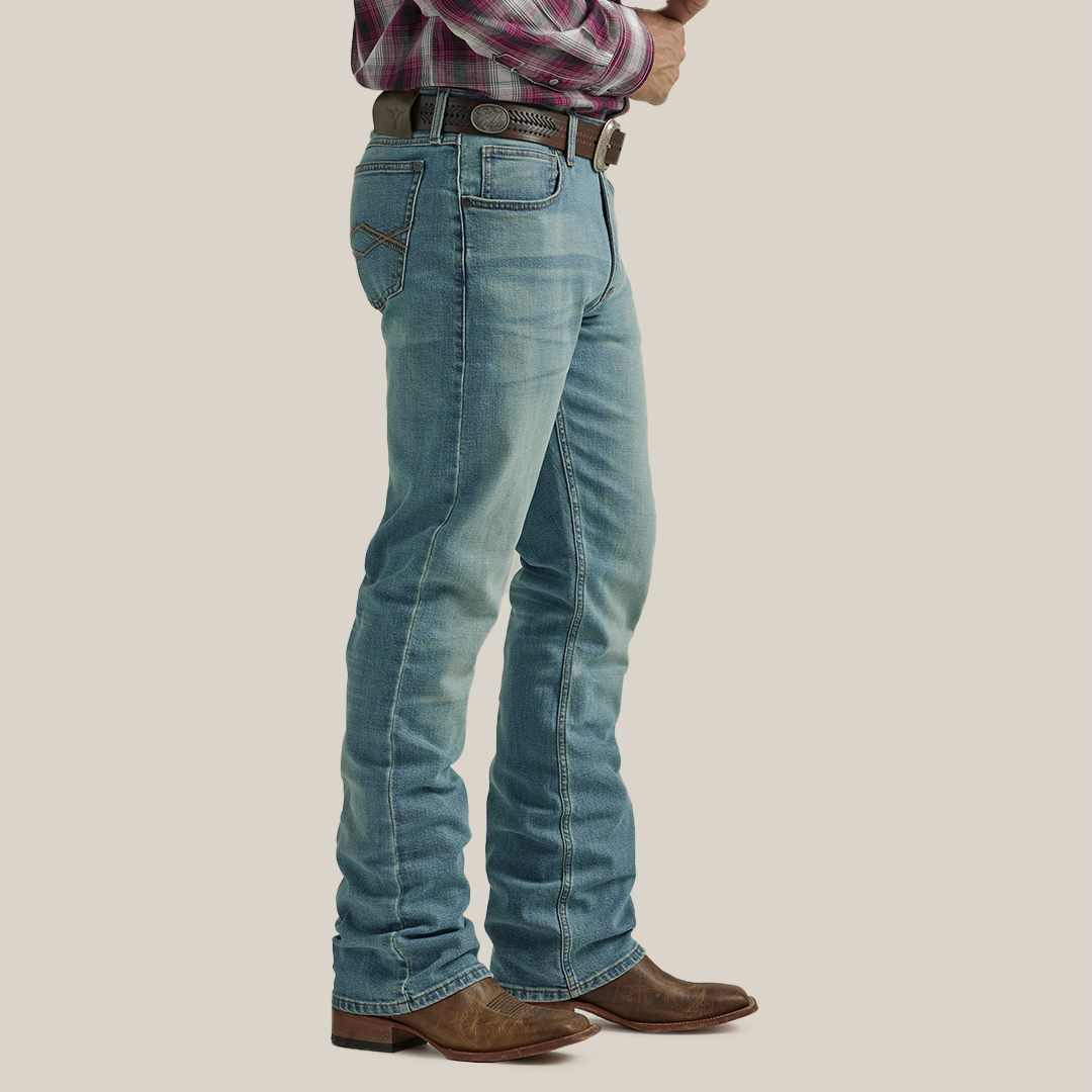 A person in Wrangler 20X Mens Light Wash Shade Bootcut Stretch Jeans - 112335407, brown leather cowboy boots, a brown belt, and a red plaid shirt stands sideways with hands clasped at their waist against a neutral background.
