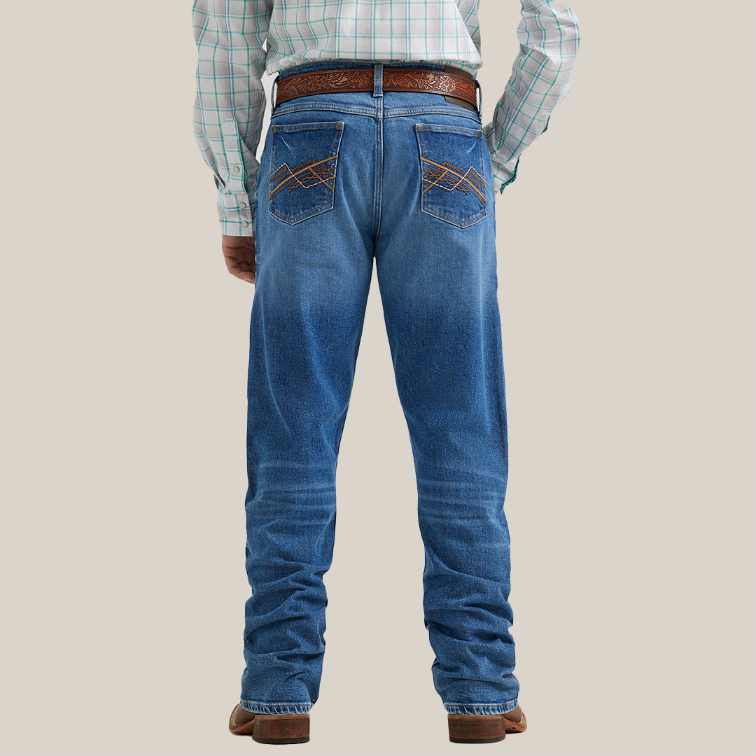 A person wearing Wrangler 20x Mens Medium Wash Backwater Vintage Bootcut Stretch Denim Jeans with a brown tooled leather belt, plaid shirt, and brown cowboy boots. The image highlights the lower half of the body against a plain background.