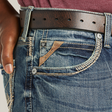 A person wearing Ariats M5 Slim Gambler Stackable Straight Leg Jeans with a brown leather belt has a hand in the pocket, showcasing contrast stitching and a leather tag on the small pocket. A partial maroon shirt is visible at the top.