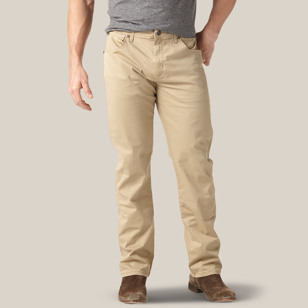 A person wearing a gray t-shirt, beige pants, and brown suede shoes stands against a neutral background. Their left hand is by their side, and their right hand rests casually in the pocket of their Mens Wrangler Retro Slim Fit Straight Leg Pant - 1088MWZFN.
