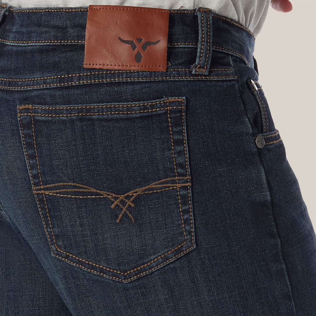 A person wears Wrangler 20X No. 33 Extreme Appleby Dark Wash Relaxed Straight Leg Jeans with dark blue premium denim, orange stitching, brown leather logo patch on the waistband, and a decorative back pocket pattern. They pair it with a crisp white shirt.