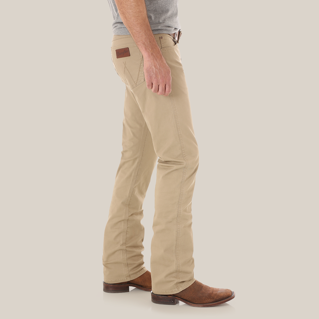 Wearing Mens Wrangler Retro Slim Fit Straight Leg Pants 1088MWZFN with a brown leather patch on the pocket, a gray shirt, and brown cowboy boots, they stand sideways against a white background, capturing the classic Wrangler Retro style.