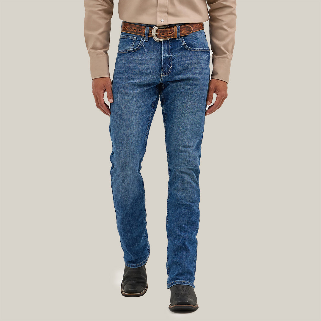 A person in a beige long-sleeve shirt, blue jeans, and a brown belt stands before a plain background. They wear Mens Wrangler 20X No. 42 Vintage Bootcut Jeans with the iconic patch, hands at their sides, while black boots complete the outfit.