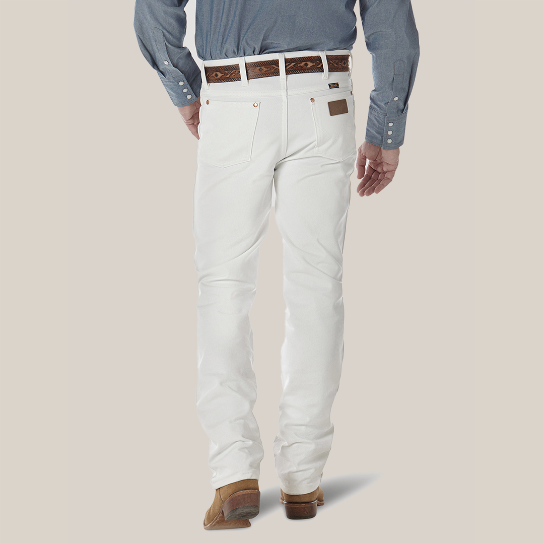 Against a light backdrop, someone is wearing Wrangler Cowboy Cut Slim Fit Jean in white, paired with a blue long-sleeve shirt, brown shoes, and a patterned brown belt. The ensemble subtly suggests a classic cowboy style.