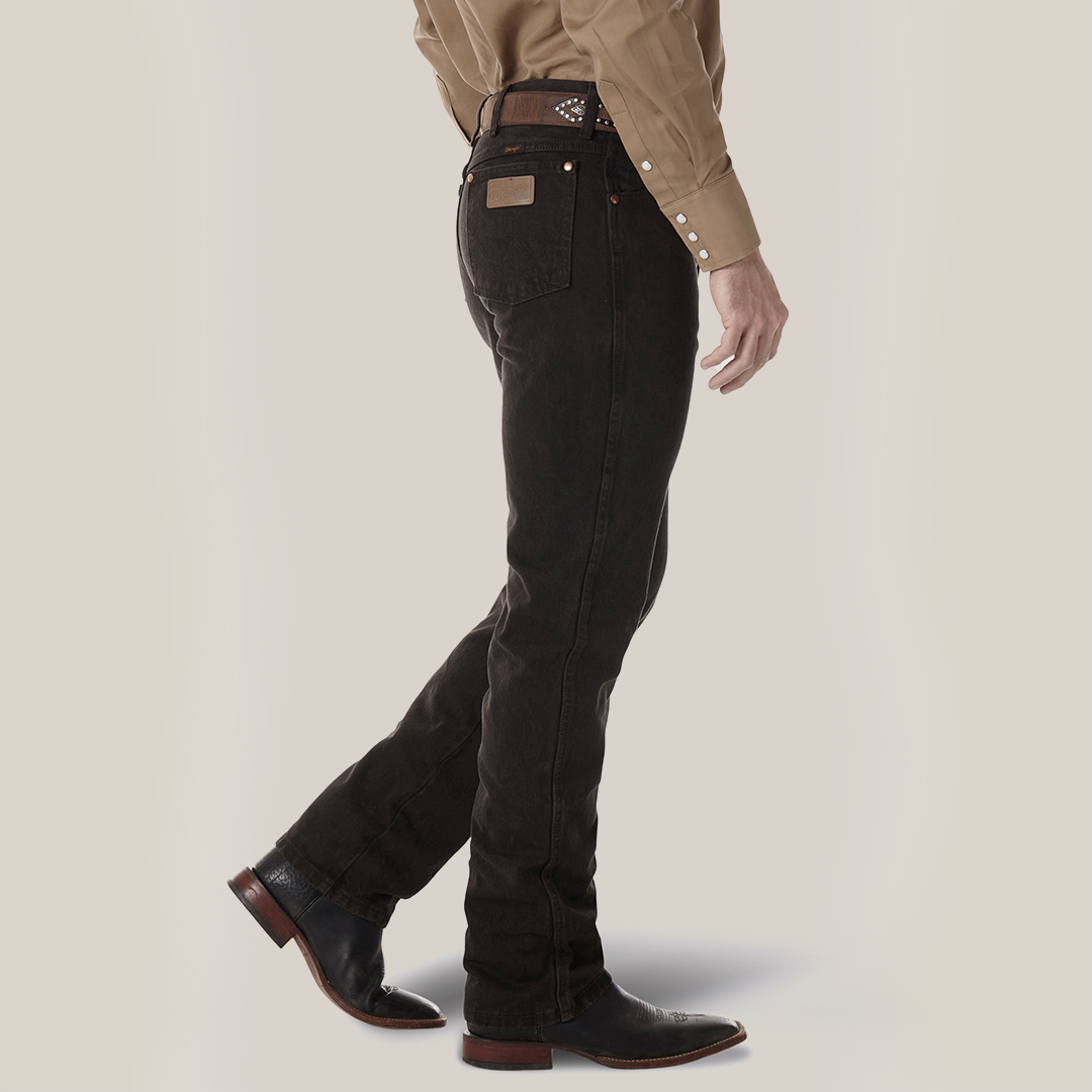 A person sporting a brown button-up shirt and dark jeans with a visible brown belt exudes rugged style. The Wrangler Cowboy Cut Slim Fit Jean - 0936KCL pairs perfectly with brown cowboy boots as they stand with one foot forward, emphasizing the side profile.