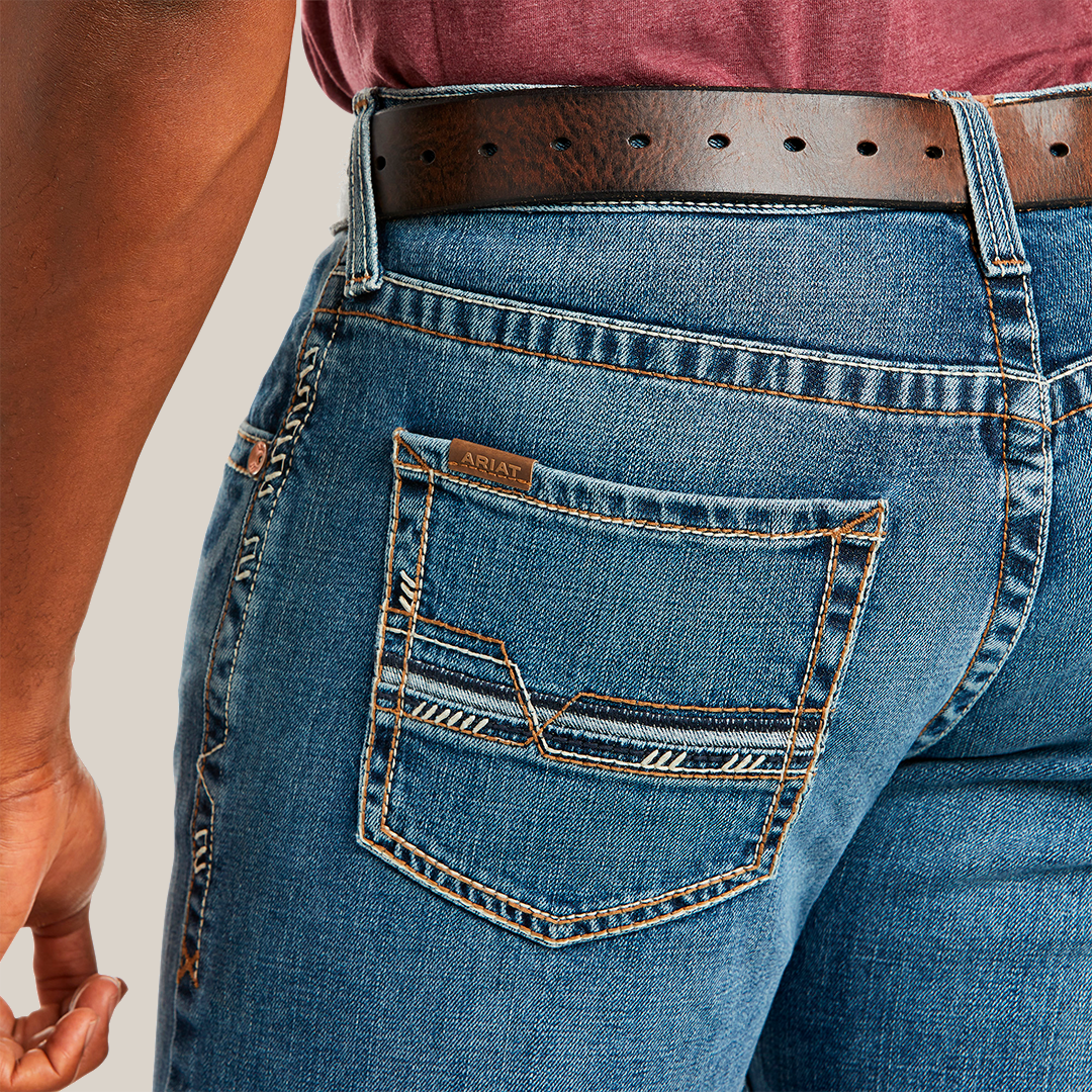 Close-up of a person in M5 Slim Stretch Stillwell Stackable Straight Leg Jean - Fargo with detailed back pocket stitching, crafted from premium cotton, secured by a brown leather belt. A maroon shirt is neatly tucked into the jeans.