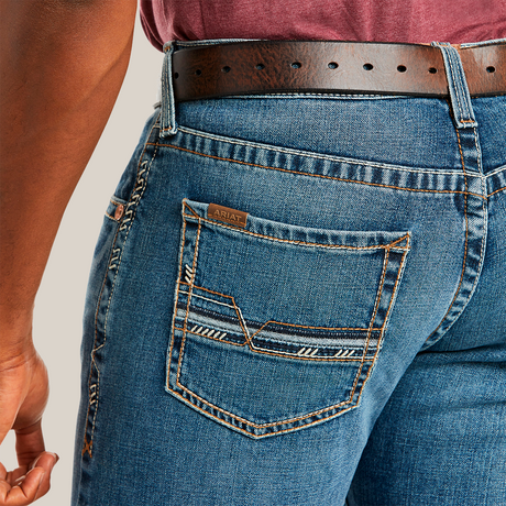 Close-up of a person in M5 Slim Stretch Stillwell Stackable Straight Leg Jean - Fargo with detailed back pocket stitching, crafted from premium cotton, secured by a brown leather belt. A maroon shirt is neatly tucked into the jeans.