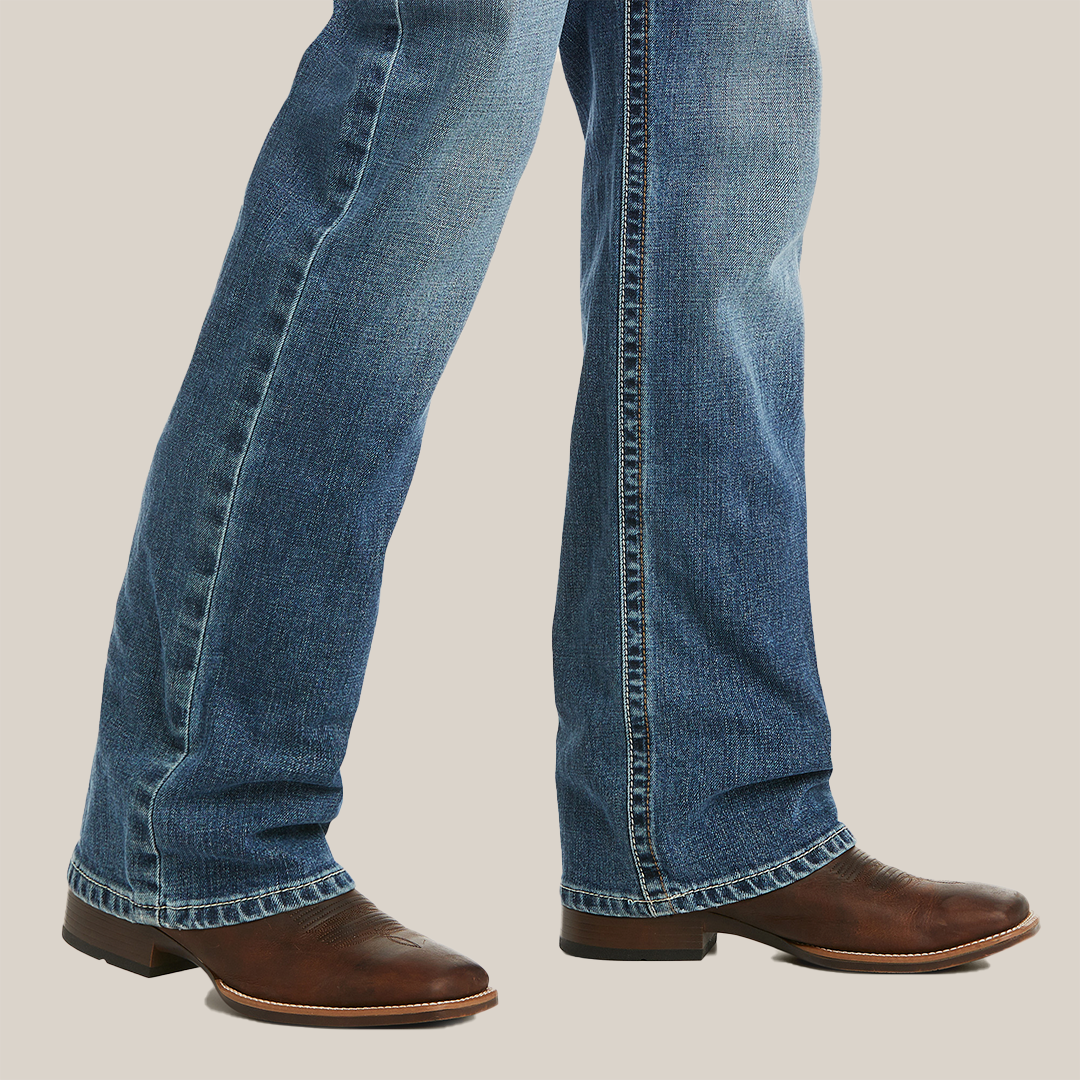 Close-up of someone wearing M5 Slim Stretch Stillwell Stackable Straight Leg Jean - Fargo over brown leather cowboy boots, slightly faded and elegantly draping over the boots against a plain, light background.