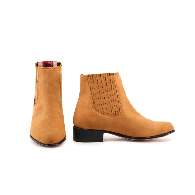 The Prime Suede Honey Round Toe boots feature tan suede, elastic side panels, black low heels, and round toes. One boot shows detailed cowboy-style stitching from the side; the other is front-facing. They have a red and plaid interior lining.