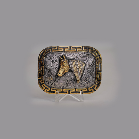 The Charro Horse Buckle features a Texas-style rectangular design with a horse head motif in rich gold tones, accentuated by swirling patterns and a Greek key border. Displayed on a clear stand against a gray backdrop, it embodies classic western designs.