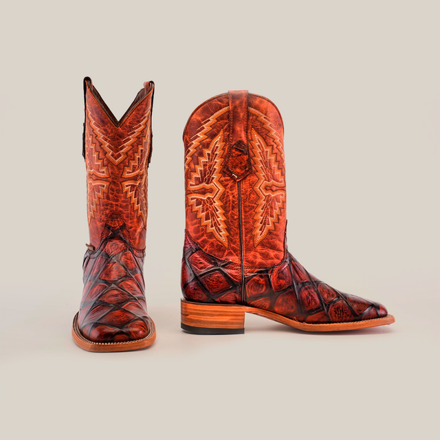 The Big Bass Pirarucu Print Cognac boots feature a square toe design. Crafted from textured brown leather, the boots are adorned with intricate patterns on the shaft and complemented by a wooden heel against a white background.