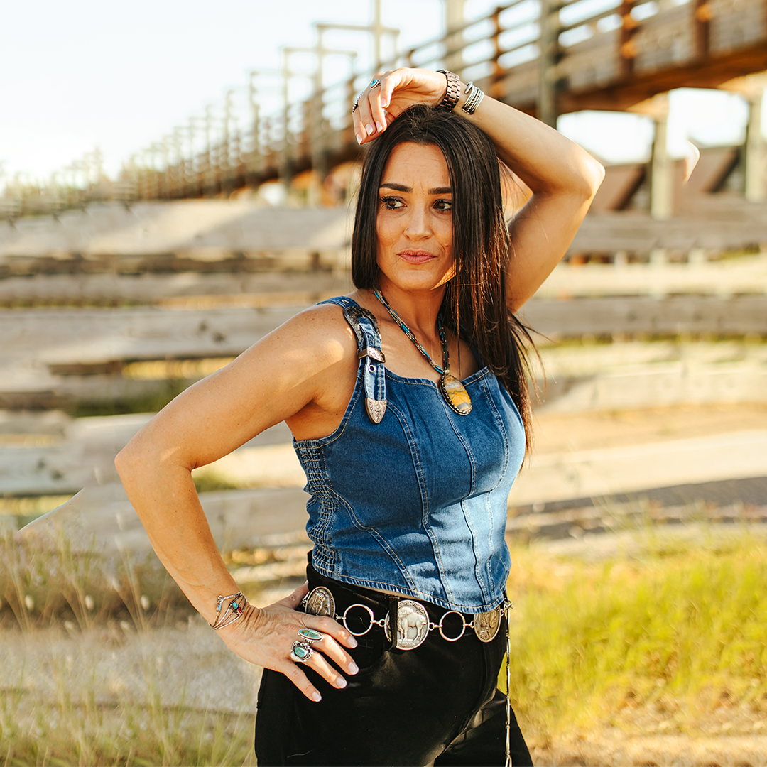 A woman with long dark hair poses confidently outdoors in the LB Denim Texas Buckle Crop, featuring a denim tank top with adjustable straps and matching black pants. Jewelry adorns her arms as she stands against wooden structures and grassy terrain under a clear sky.