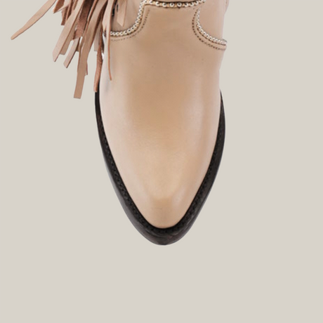 A close-up of a Praga Fringe Tall Shaft Cream - J Toe boot, showcasing decorative stitching and fringe details on the beige leather shaft. The pointed toe has a black edge, all set against a plain white background.