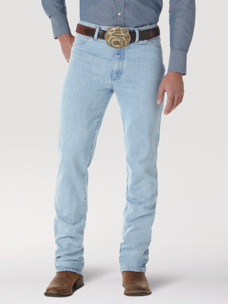 A person in Wrangler Cowboy Cut Slim Fit Jeans, wearing a brown belt with an ornate buckle, blue shirt, and brown boots. The premium denim fits perfectly against a plain light background, although their head is not visible.