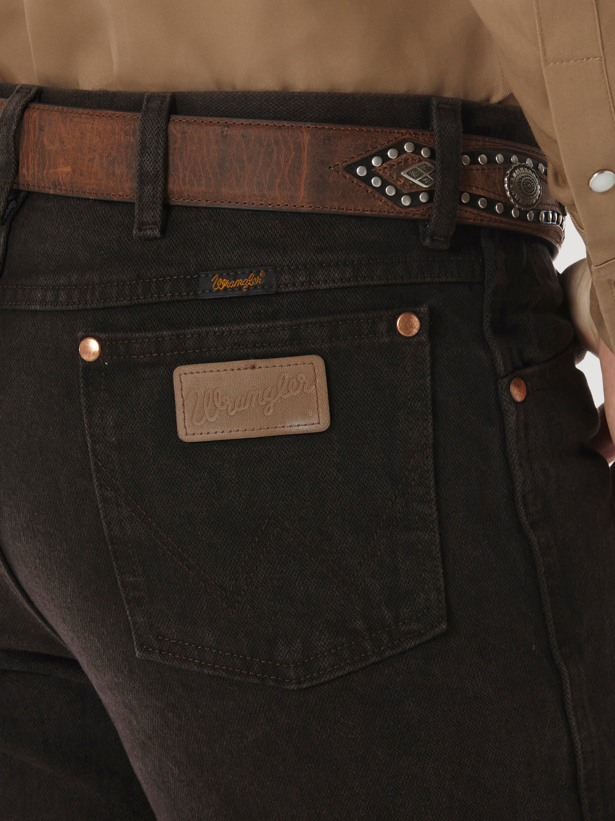 Close-up of a person in Wrangler Cowboy Cut Slim Fit Jean - 0936KCL, dark denim with a leather belt featuring silver details. The tan shirt is partially visible, but the iconic Wrangler label on the back pocket remains the focus.