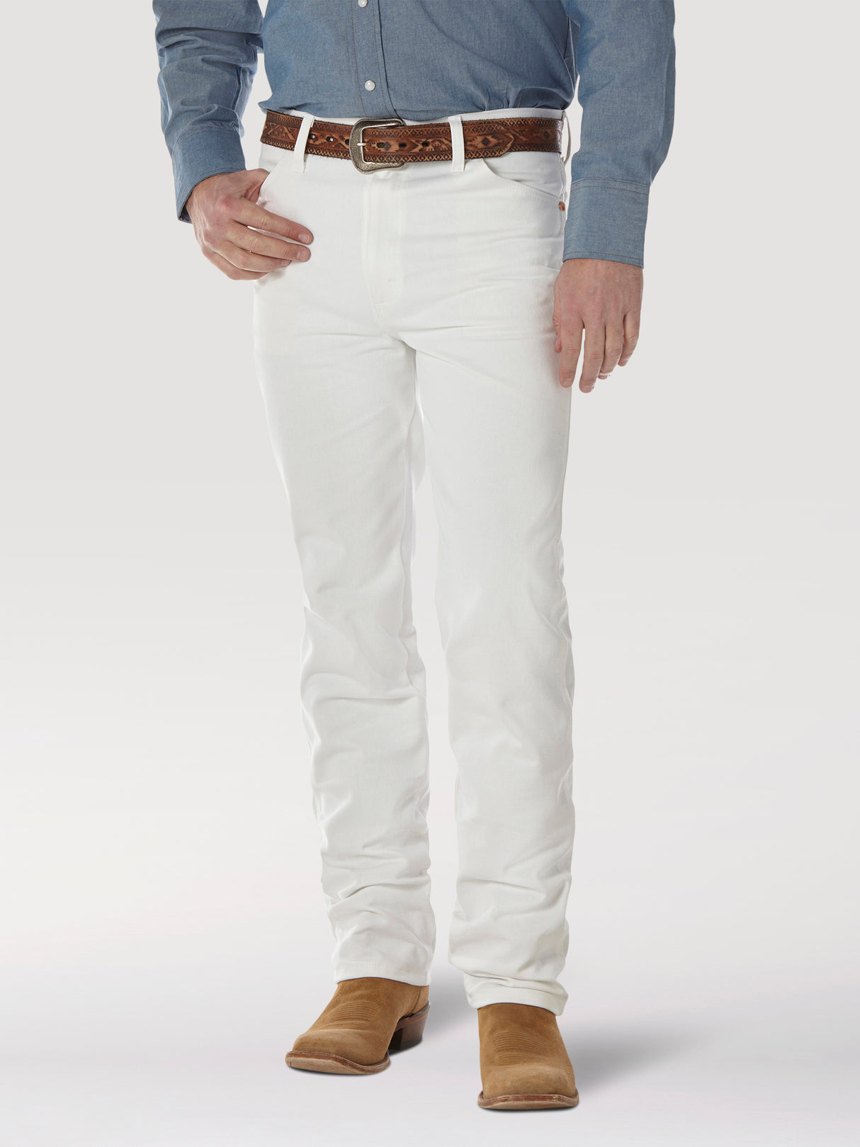 A person wearing a blue button-up shirt, Wrangler Cowboy Cut Slim Fit Jeans (0936WHI), and a brown leather belt is grounded in brown suede shoes. The iconic Wrangler patch enhances the look against the plain white background.