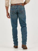 A person in Wrangler Mens 20X Extreme Relaxed Fit Jeans - 1044MWXMA, blue denim with a brown belt and shoes, stands against a white background. They sport an arm tattoo and light-colored rolled sleeves, epitomizing the relaxed style.