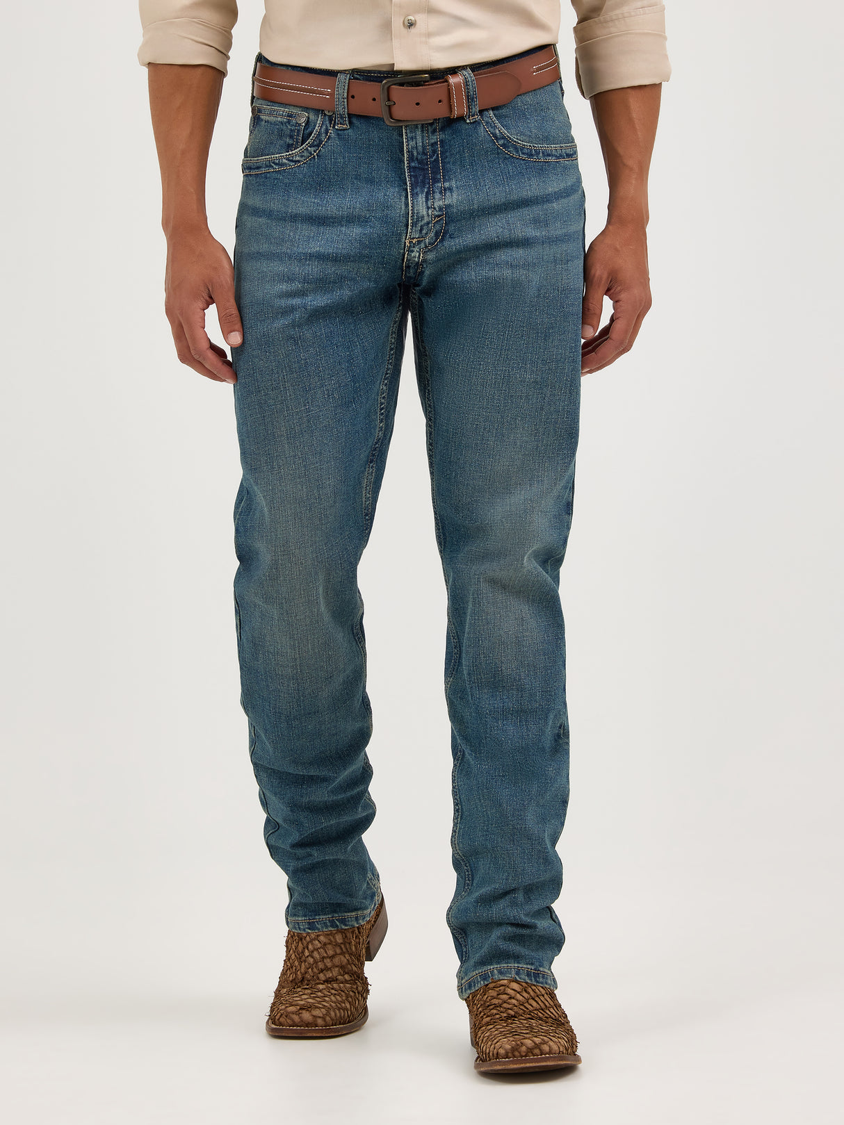 The lower half of a person is visible, wearing Wrangler Mens 20X Extreme Relaxed Fit Jeans - 1044MWXMA in Premium Denim blue with a worn look. The outfit is completed with a beige shirt, brown leather belt, and woven brown shoes against a plain background.