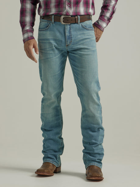 Someone in a plaid shirt, Wrangler 20X Mens Light Wash Shade Bootcut Stretch Jeans, and brown cowboy boots stands against a plain background. The light blue jeans are slightly faded and secured with a brown belt featuring an oval buckle.