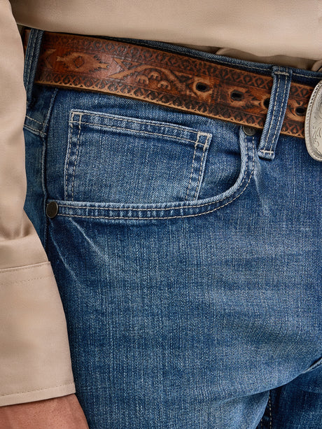 Close-up of a person wearing Mens Wrangler 20X No. 42 Vintage Bootcut Jean in premium denim blue, paired with a patterned brown leather belt featuring a decorative metal buckle and a portion of a light-colored shirt visible on the left.