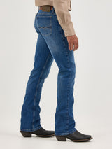 A person in blue denim jeans and black boots is shown from the side against a plain background, wearing a beige shirt with white buttons. The jeans, Mens Wrangler 20X No. 42 Vintage Bootcut Jean - 112338537, feature stitching details and a distinctive Wrangler patch on the back pockets.