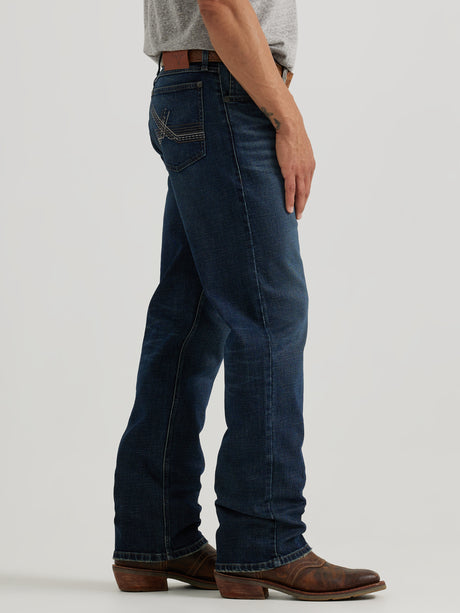 A man in a gray t-shirt and leather belt stands sideways wearing dark blue Wrangler 20X Sunnybrook Dark Wash Extreme Relaxed Straight Stretch Denim Jeans, featuring visible back pocket stitching, and brown cowboy boots. The background is plain gray.