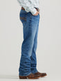 A person wearing Wrangler 20x Mens Medium Wash Backwater Vintage Bootcut Stretch Denim Jeans, a plaid shirt, and brown cowboy boots stands in profile with hands in pockets against a neutral background.