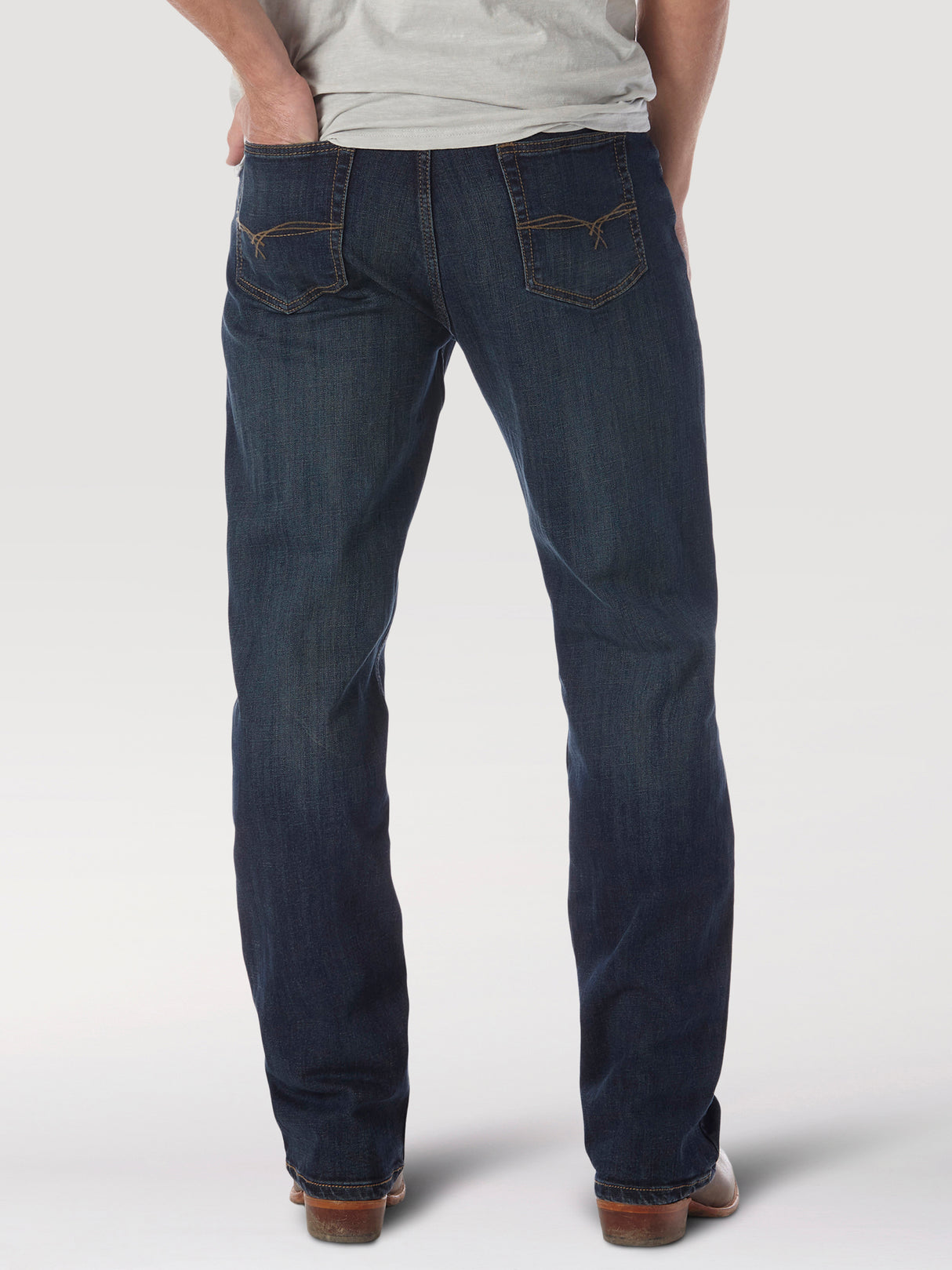 A person poses against a white backdrop wearing the Wrangler 20X No. 33 Extreme Appleby Dark Wash Relaxed Straight Leg Jeans, featuring orange stitching and styled with brown shoes, embodying a laid-back essence with hands in back pockets and a light gray shirt.