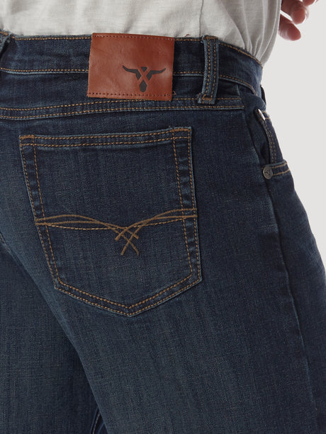 A person wears Wrangler 20X No. 33 Extreme Appleby Dark Wash Relaxed Straight Leg Jeans with dark blue premium denim, orange stitching, brown leather logo patch on the waistband, and a decorative back pocket pattern. They pair it with a crisp white shirt.