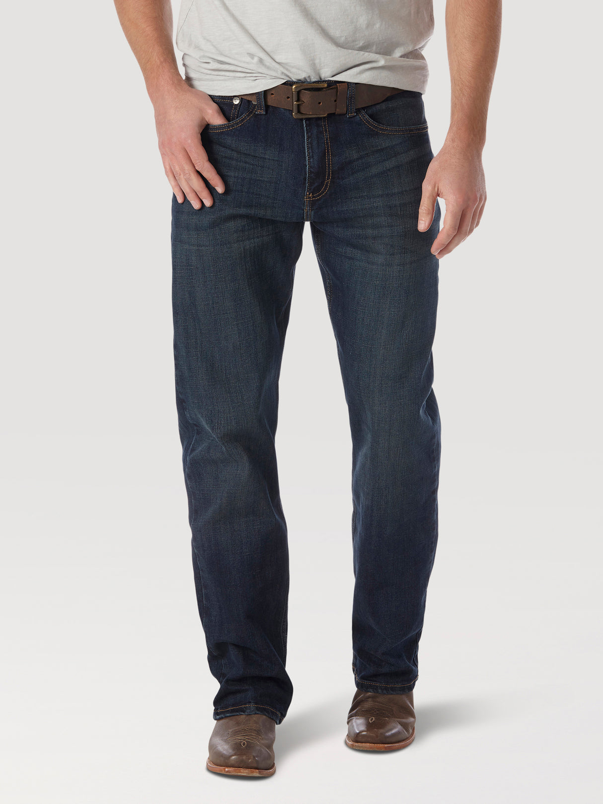 A person wearing a gray t-shirt, Wrangler 20X No. 33 Extreme Appleby Dark Wash Relaxed Straight Leg Jeans - 1033MWXAB, and brown leather shoes stands on a white background. Their right hand is in a pocket, highlighting the premium denim style with a coordinating brown belt.