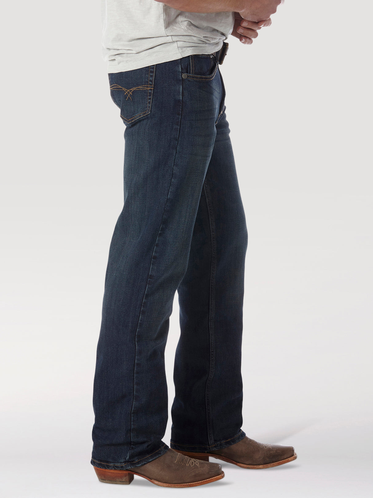 A person wearing Wrangler 20X No. 33 Extreme Appleby Dark Wash Relaxed Straight Leg Jeans and brown cowboy boots, hands on their waist. The jeans relaxed fit blends effortlessly with the plain light gray background.