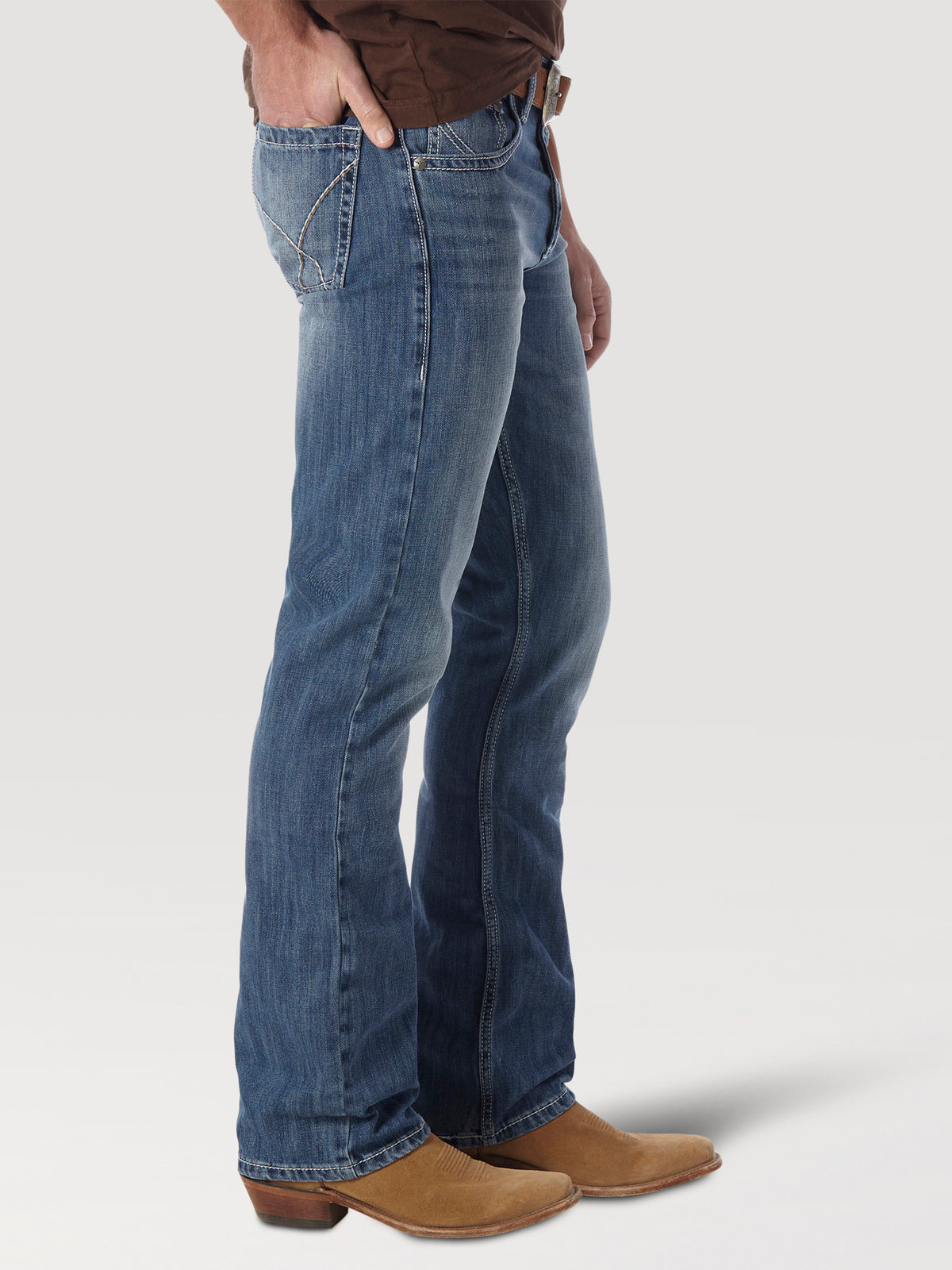 A person, shown from shoulders down, wears a brown shirt, premium denim blue jeans with decorative back pocket stitching, and Mens Wrangler 20X No. 42 Vintage Bootcut Jeans. They stand against a plain light gray background.