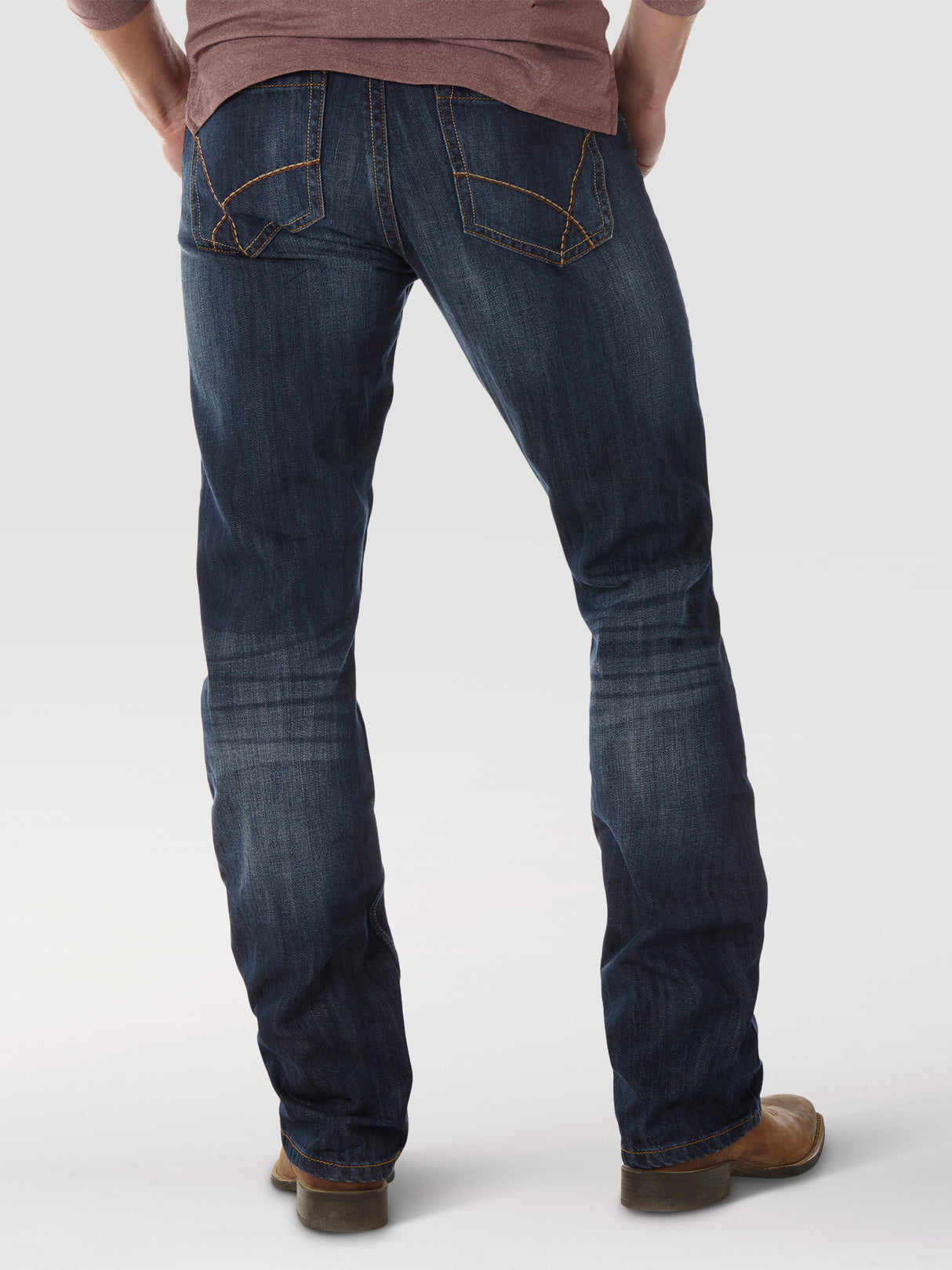 The image shows a person from behind wearing Wrangler® 20X® No. 42 Vintage Boot Jeans in dark blue with brown stitching and brown shoes, paired with a maroon long-sleeve top, all set against a light gray background.