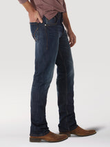 A person wearing Wrangler® 20X® No. 42 - Vintage Boot Jean 1042MWXRD, dark blue premium denim jeans, stands sideways with brown boots and one hand in their pocket. The vintage bootcut jeans have a slight fade and classic fit, paired perfectly with a long-sleeved reddish-brown top.