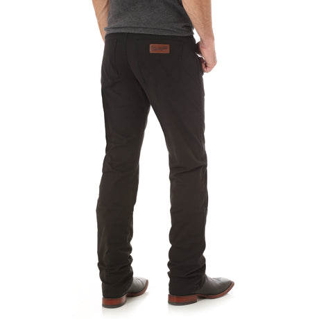 A person in Mens Wrangler Retro Slim Fit Straight Leg Pant - 88MWZBK and sleek black shoes stands facing away. These premium denim pants with a leather patch are paired with a dark gray shirt on a plain white background, showcasing a relaxed yet sophisticated style.