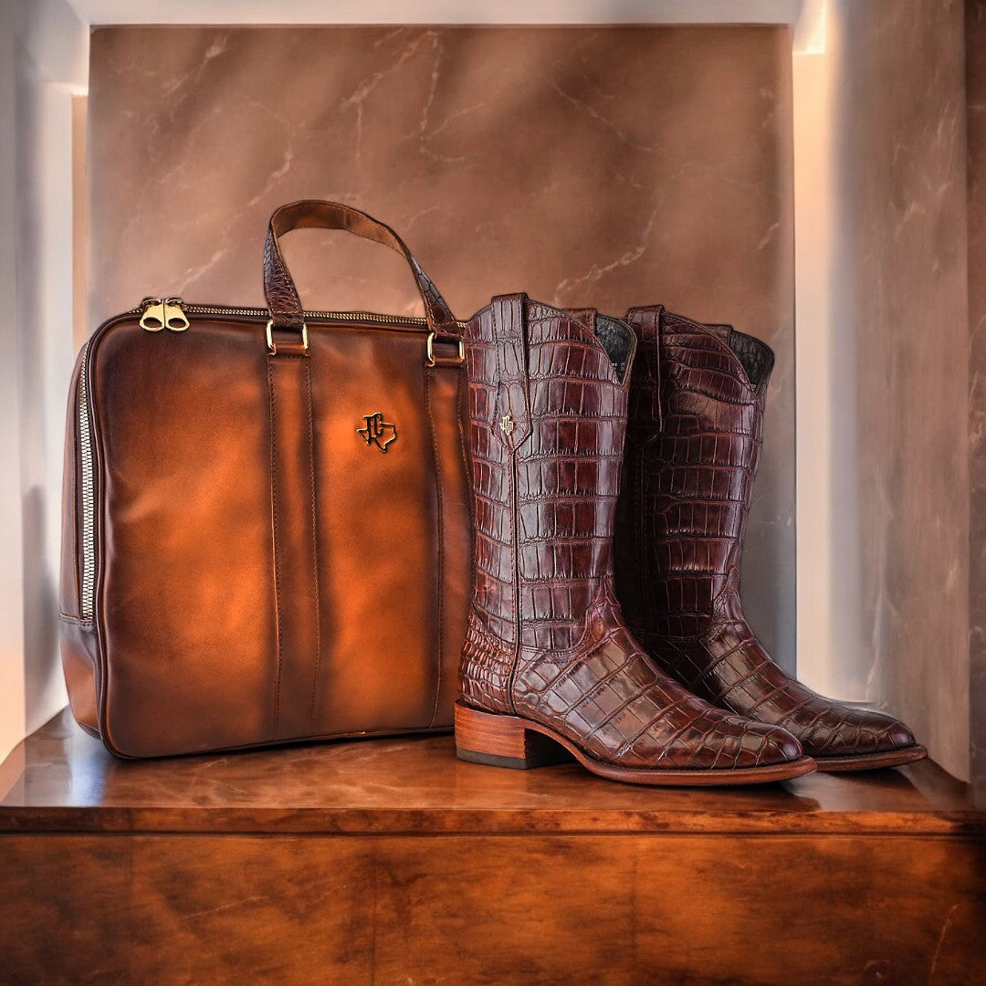 On a wooden shelf with a marble backdrop, an exquisite pair of Exotic Full American Alligator boots in brown complements a matching leather briefcase adorned with a sleek logo.