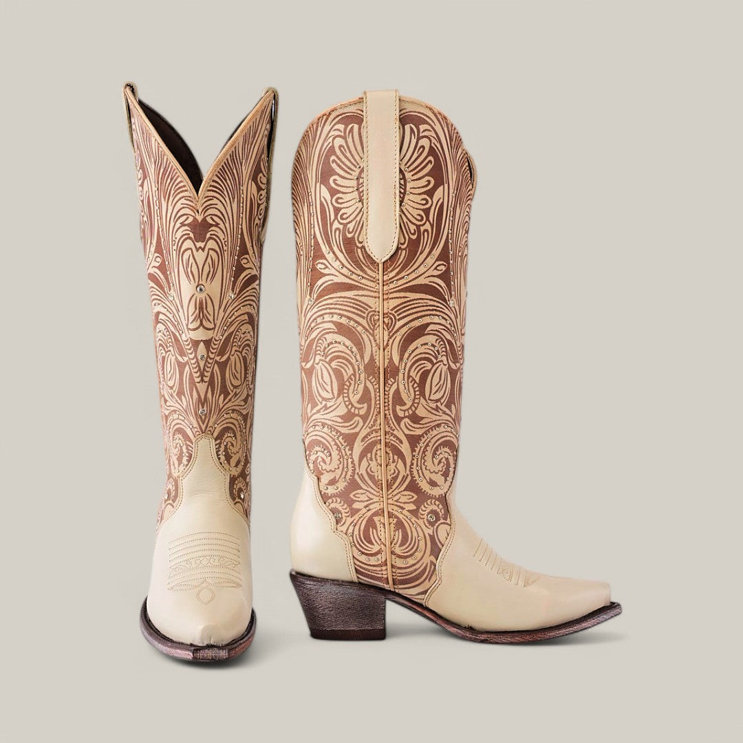 A pair of Cavalier Laser Cream boots features intricate floral embroidery on a tall shaft, with one boot upright and the other angled. They showcase a snip toe design, stacked heels, and a light gray background.