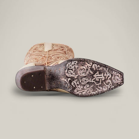 The image displays a Cavalier Laser Cream - Tall Shaft - Snip Toe boot, positioned with the sole up to highlight floral engravings and a logo. Its tan upper boasts decorative patterns against a neutral gray background, exuding elegance.