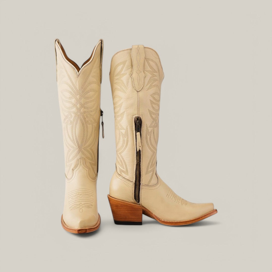 The Kellie Bone Leather Sole boots feature a tall shaft, intricate embroidery, snip toe design, wooden heels, side zipper with writing, and decorative stitching. One boot stands upright while the other is angled to highlight its snip toe and detailed craftsmanship.