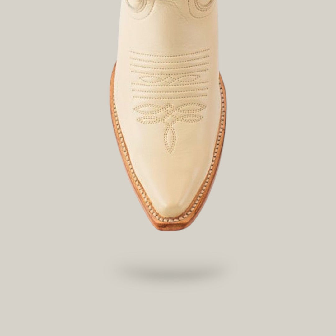 The Kellie Bone Leather Sole boot features a cream color with intricate stitching and boasts a snip toe, set against a light gray background. Its tall shaft is adorned with detailed floral and linear embroidery, complemented by a brown sole and defined edge.